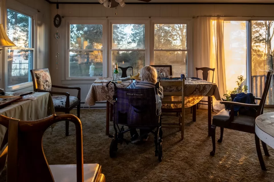 Senior woman sitting inchair looking out large window at golden hour in Broomes Island, Maryland.